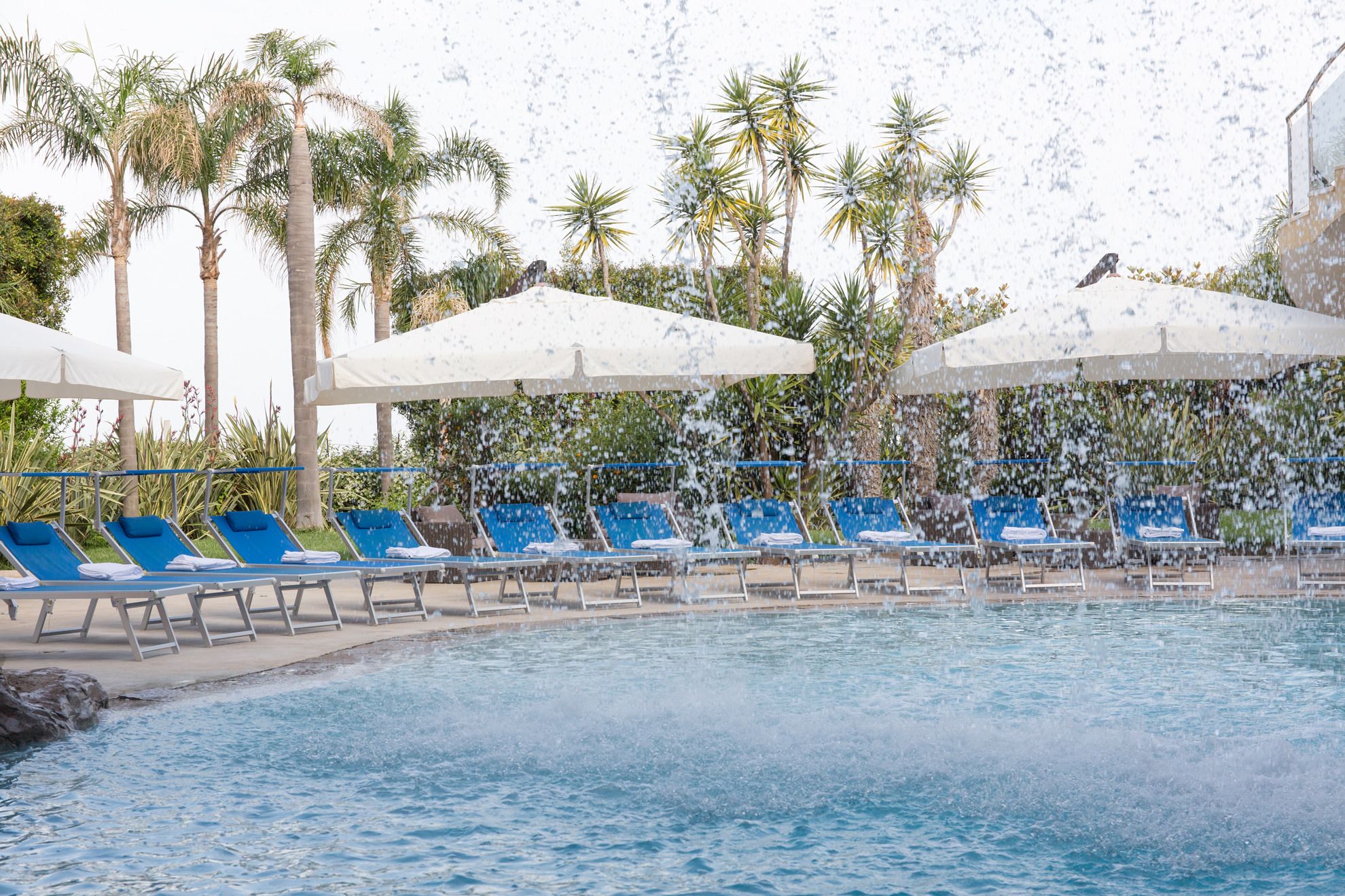 A poolside area with blue lounge chairs and white umbrellas, surrounded by palm trees.