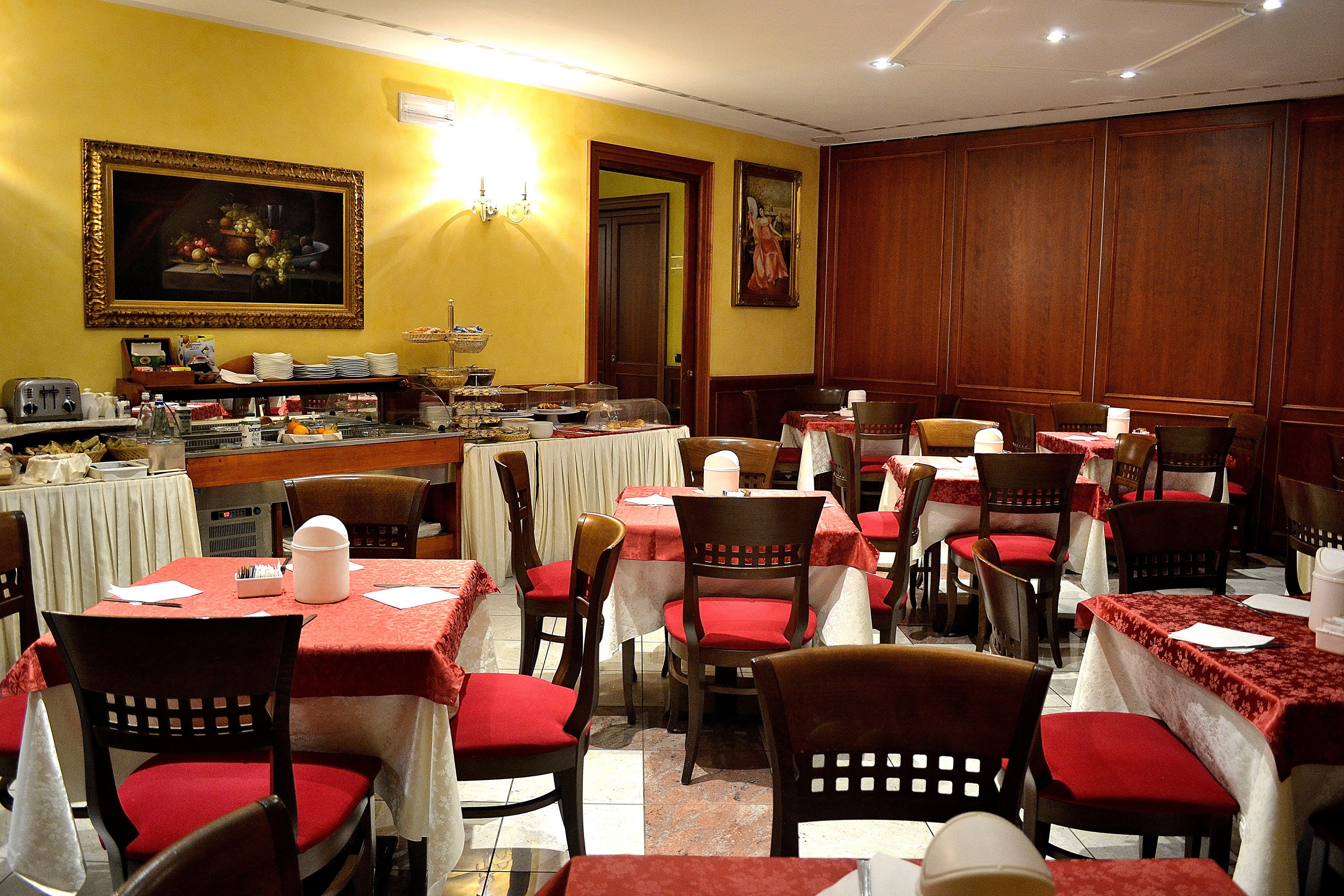 Uma área de refeições com mesas preparadas para o serviço, cadeiras com almofadas vermelhas e um buffet com diversos alimentos.