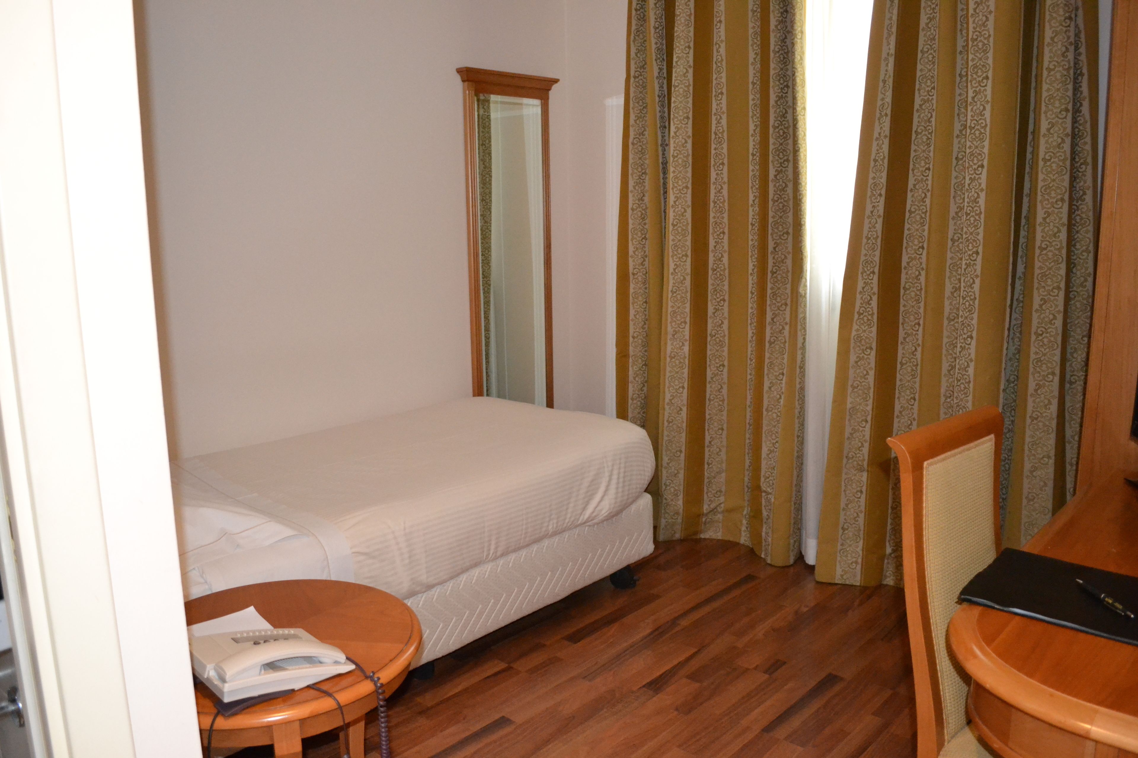 A small, neatly arranged bedroom with a single bed, a wooden chair, a small table, and a mirror.