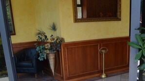 Een hotelentree met een betegelde vloer, een zwarte stoel, een potplant, een spiegel en een messing vloerlamp.
