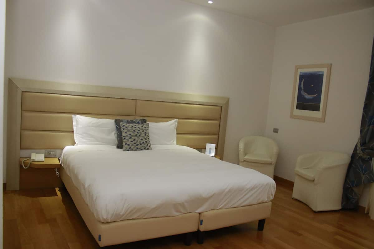 A spacious bedroom with a large bed, a bedside table with a phone, a chair, and a framed picture on the wall.