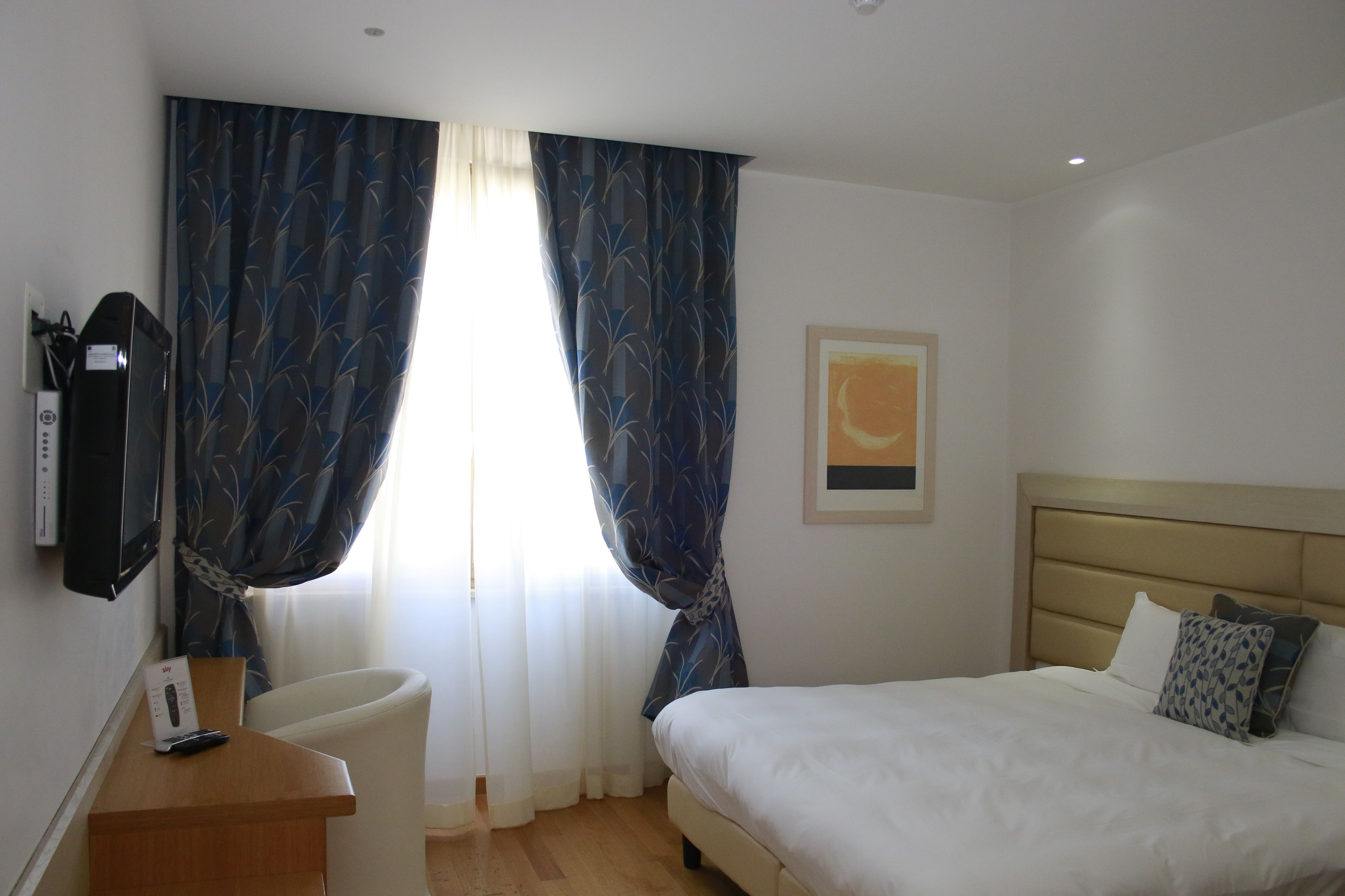 A hotel room with a bed, a desk with a chair, a television mounted on the wall, and a window with curtains.