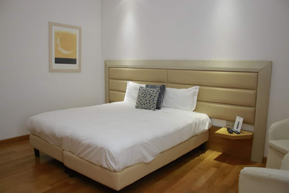 A neatly made bed with a headboard, a bedside table with a phone and remote, and a chair to the right.