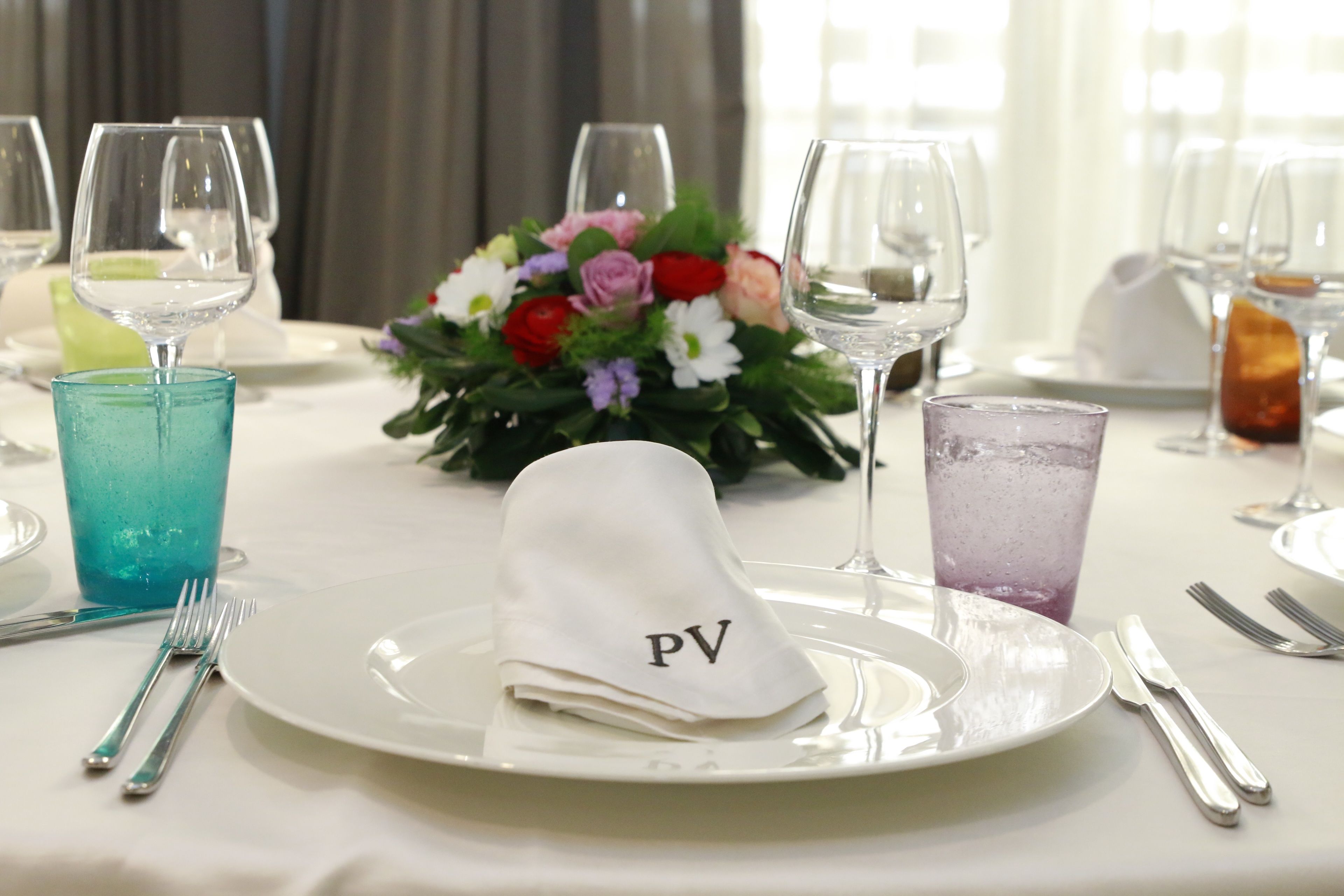 Ett bord dukat med en vit tallrik, en vikt servett med bokstäverna ”PV” på, ett blått glas, ett lila glas och en blomsterdekoration i mitten.