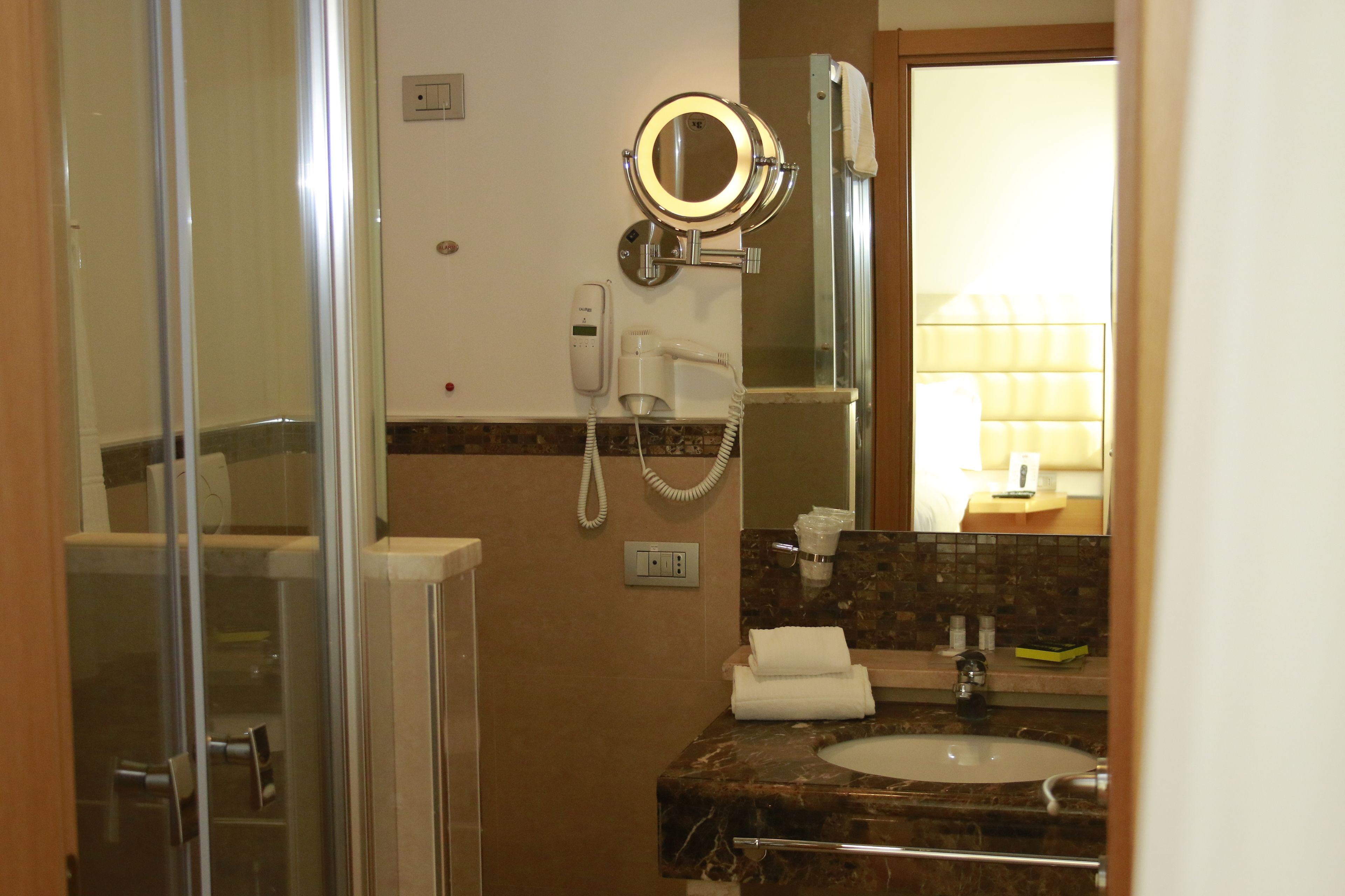 Une salle de bain dotée d’une douche à parois de verre, d’un lavabo avec un plan de toilette en marbre, d’un miroir à cadre rond et d’un sèche-cheveux.