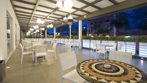 An indoor seating area with white chairs and tables, a patterned table in the foreground, and a view of a building with palm trees in the background. - Palazzo Virgilio (Brindisi)