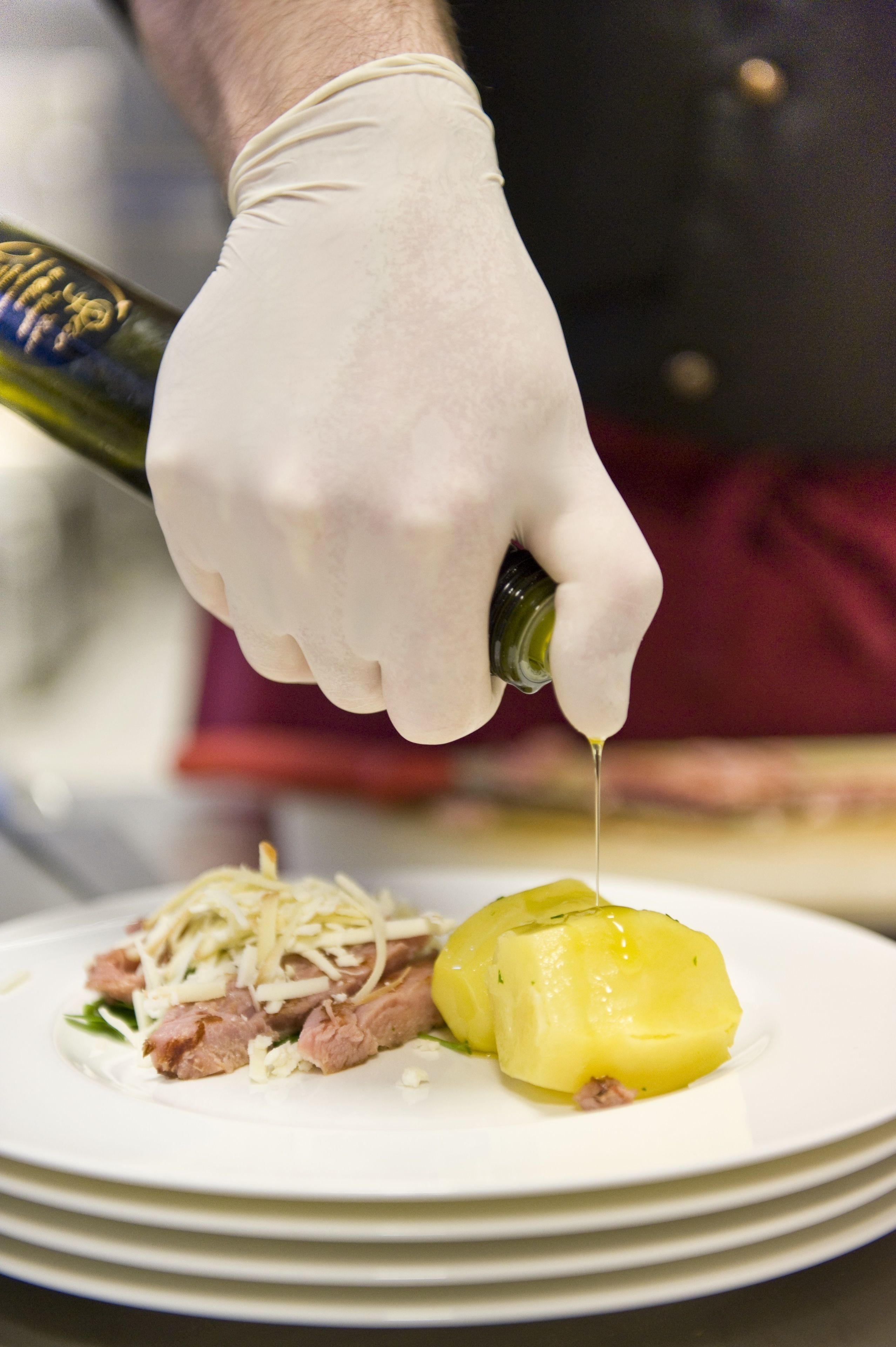 Eine Person in einem weißen Handschuh gibt Olivenöl über ein Gericht mit Fleisch und Kartoffeln.