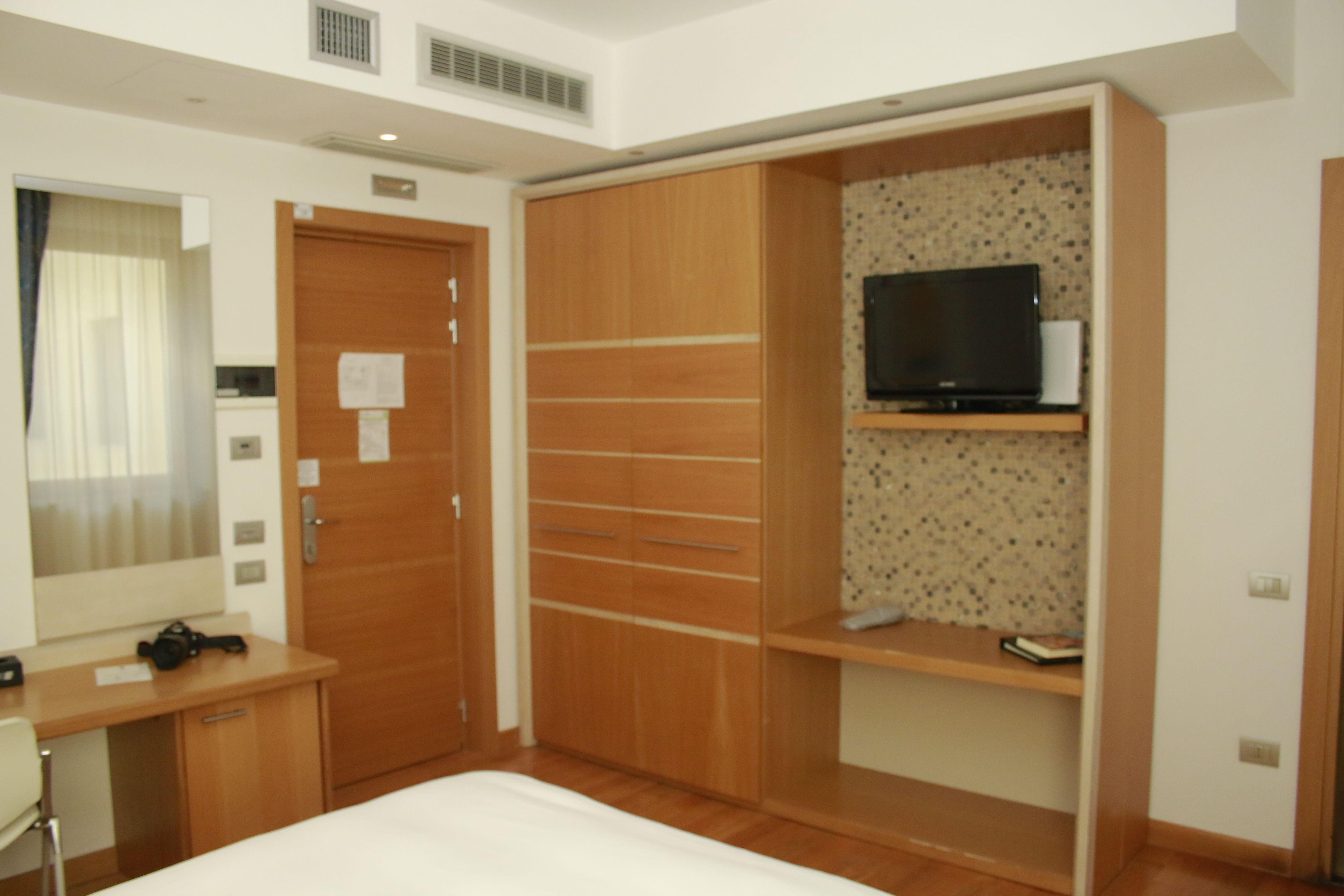 A hotel room with a bed, a wooden wardrobe, a desk with a chair, a television on a shelf, and a mirror.