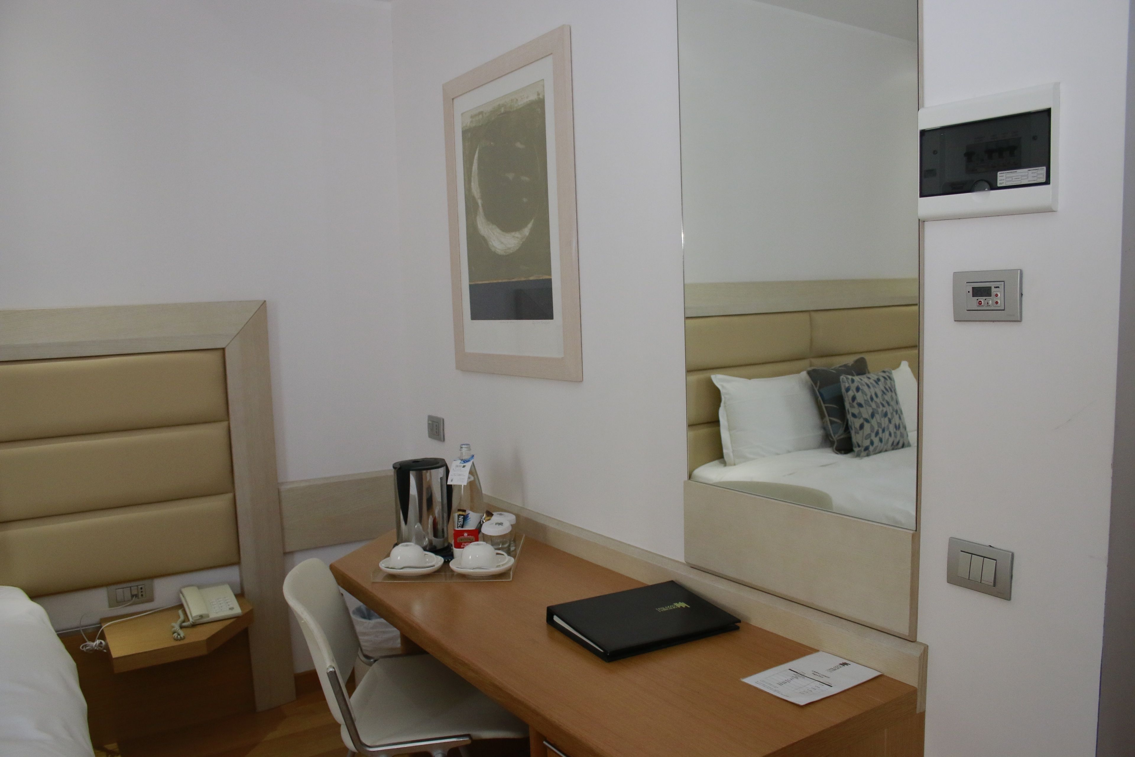 A hotel room with a bed, a desk with a chair, a coffee maker, a phone, and a mirror reflecting the bed.