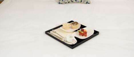 Una cama bien hecha con sábanas blancas, una bandeja con alimentos para el desayuno y una almohada estampada.