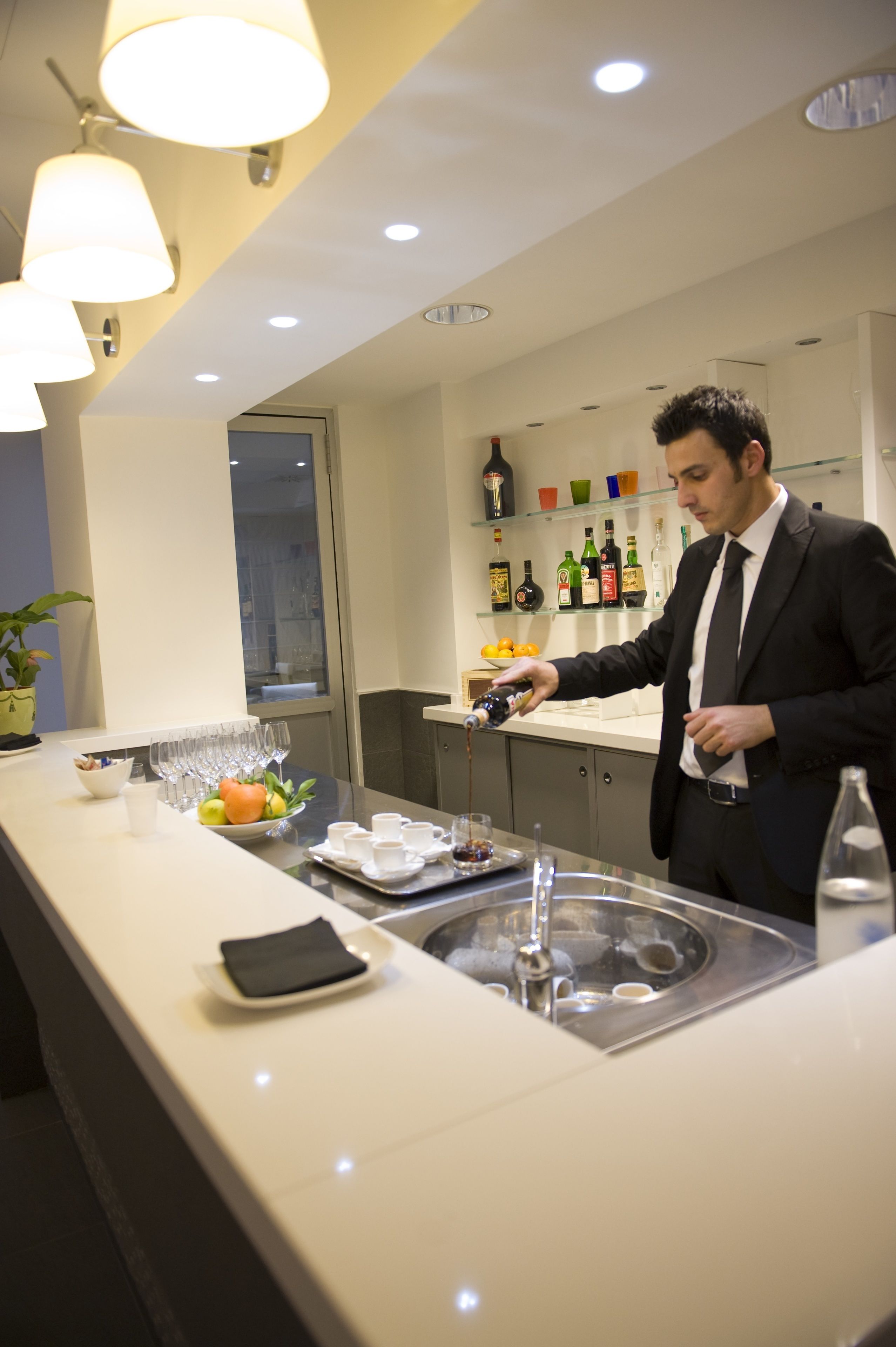 A man in a suit pouring a drink into a glass at a bar with a white countertop, glasses, and bottles in the background.