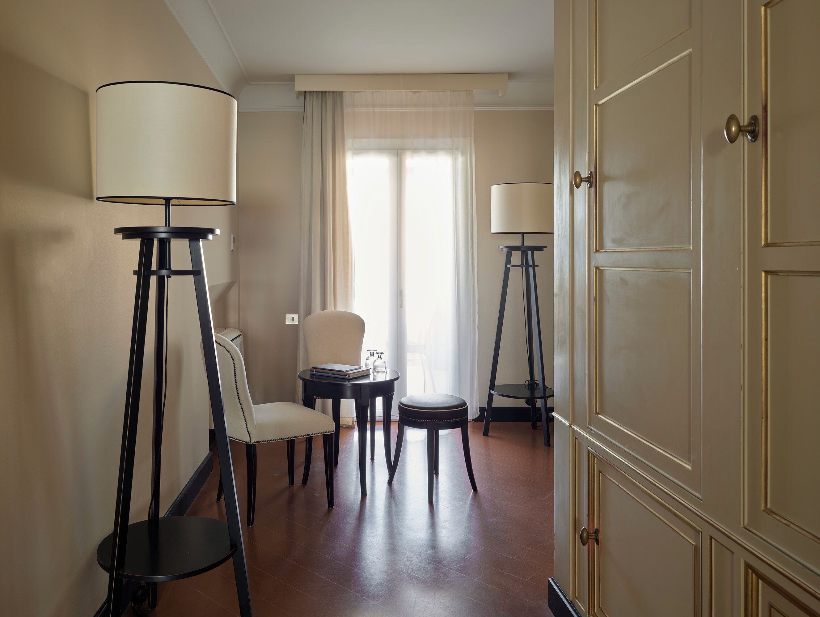 A hotel room with a dining area, two standing lamps, a chair, a small table with books, and a door.