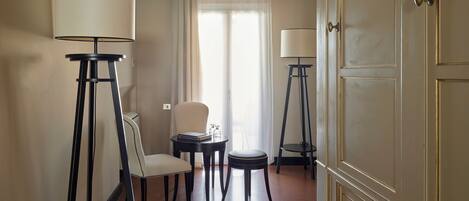 Habitación de hotel con comedor, dos lámparas de pie, una silla, una pequeña mesa con libros y una puerta.