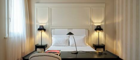 A hotel room with a bed, two bedside lamps, a desk with a chair, and a window with sheer curtains.