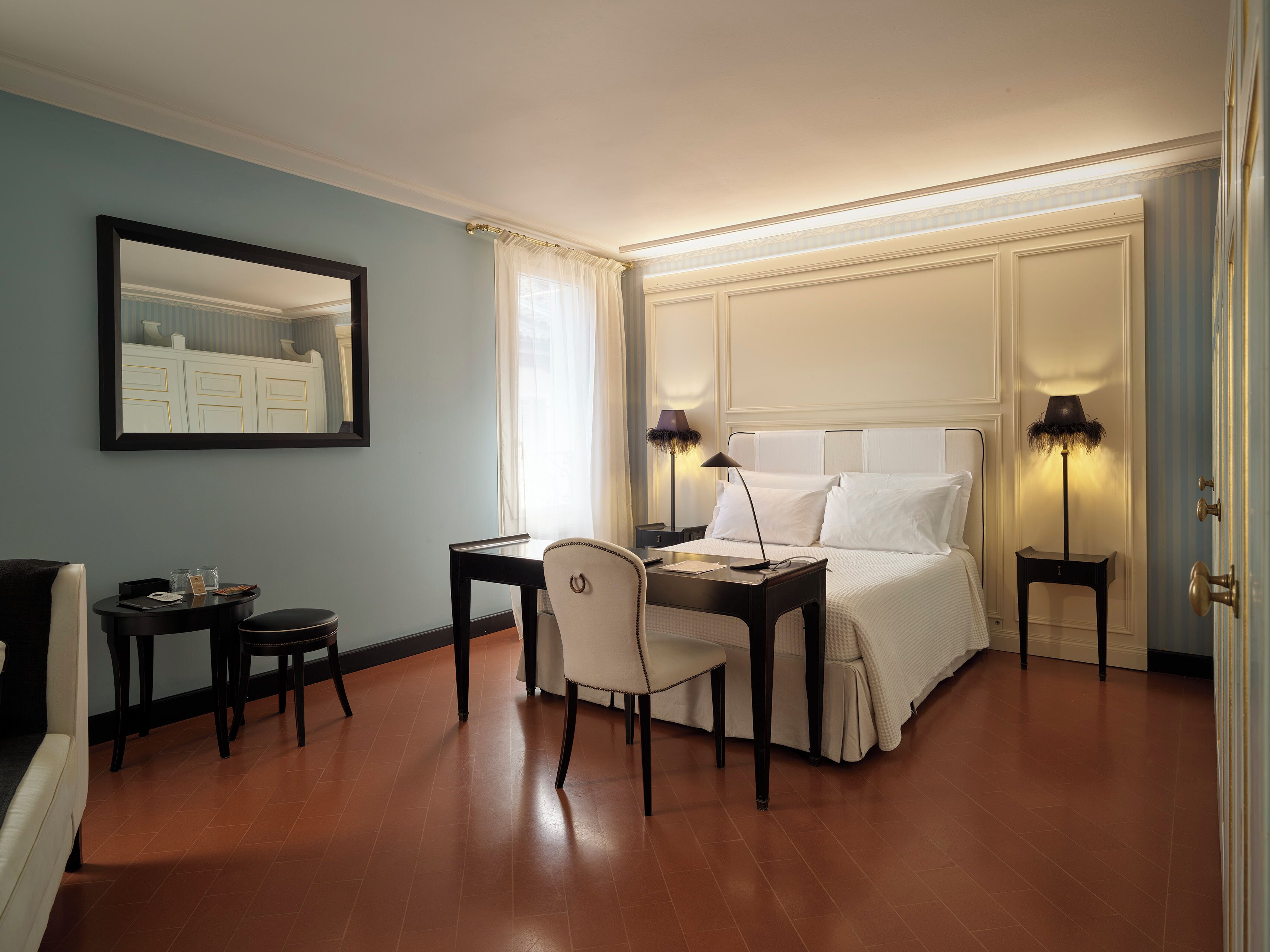 Une chambre d’hôtel comprenant un lit, deux tables de chevet, un bureau avec une chaise, un miroir et une fenêtre avec des rideaux.