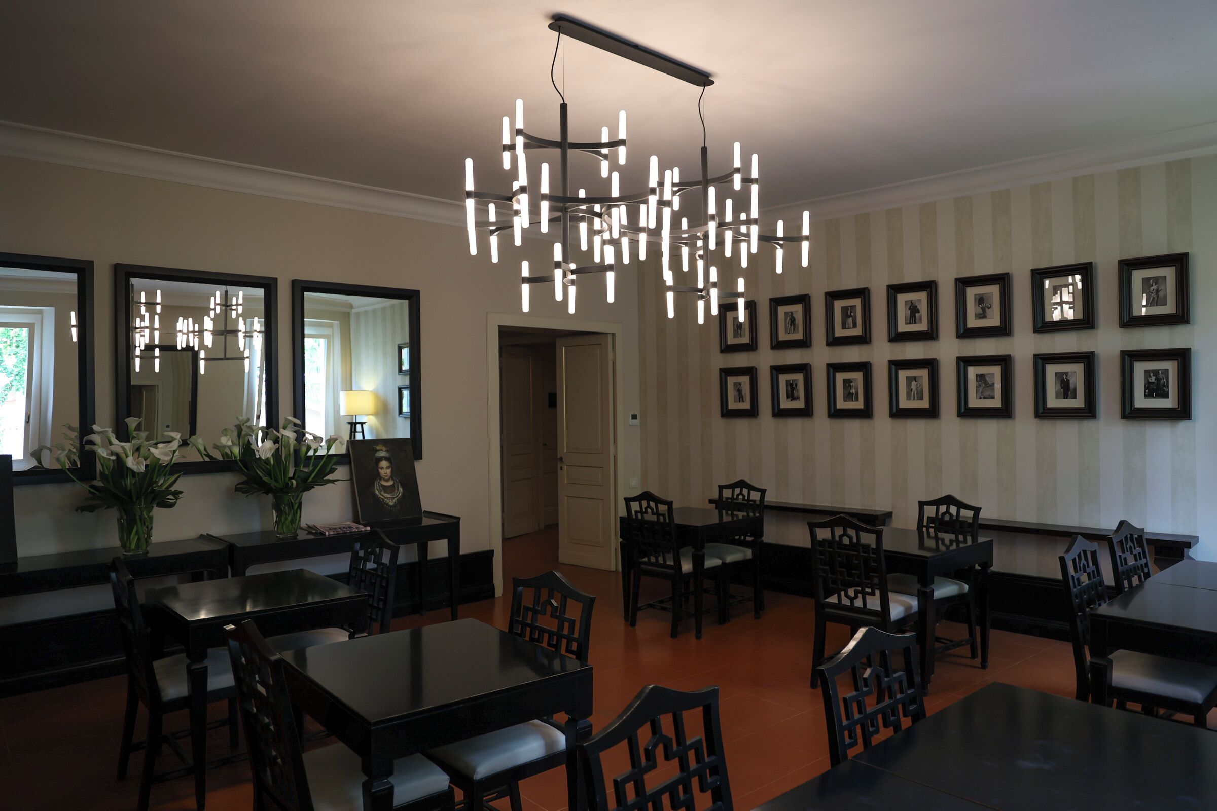 A dining area with multiple tables and chairs, a chandelier, framed pictures on the wall, and a mirror reflecting the room.