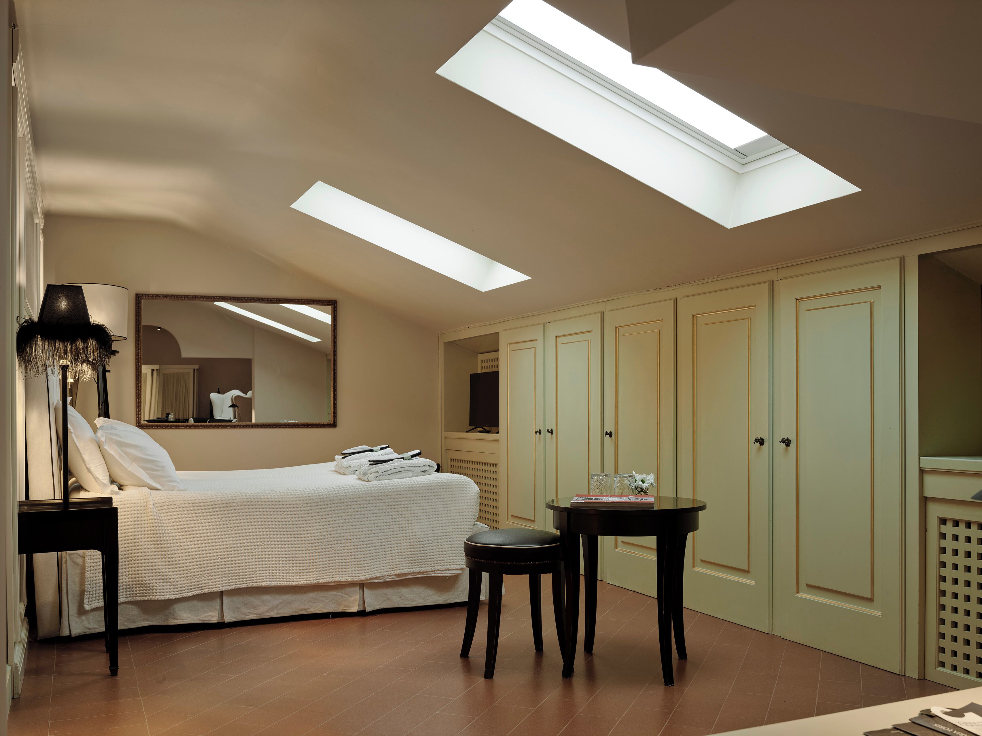 A bedroom with a skylight, a bed, a nightstand, a chair, a table, and a wardrobe.