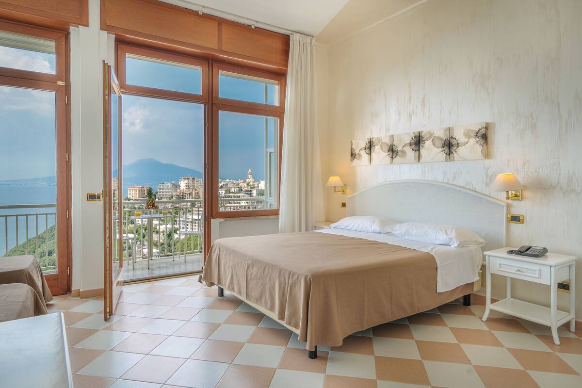 A hotel room with a bed, a nightstand, a view of the city, and a balcony.