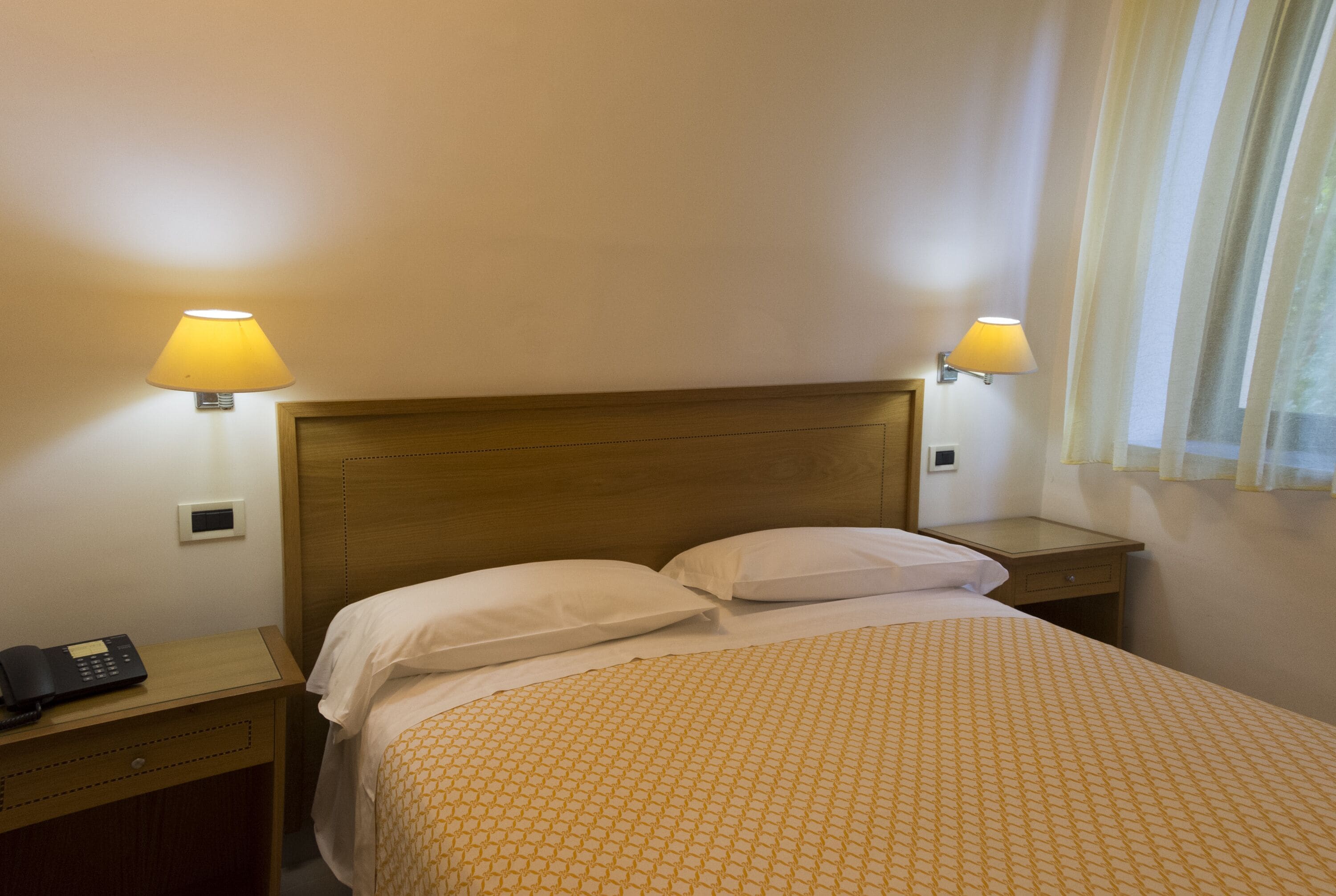 Quarto de hotel com cabeceira de madeira, duas luminárias de cabeceira, telefone na mesinha de cabeceira e colcha estampada.