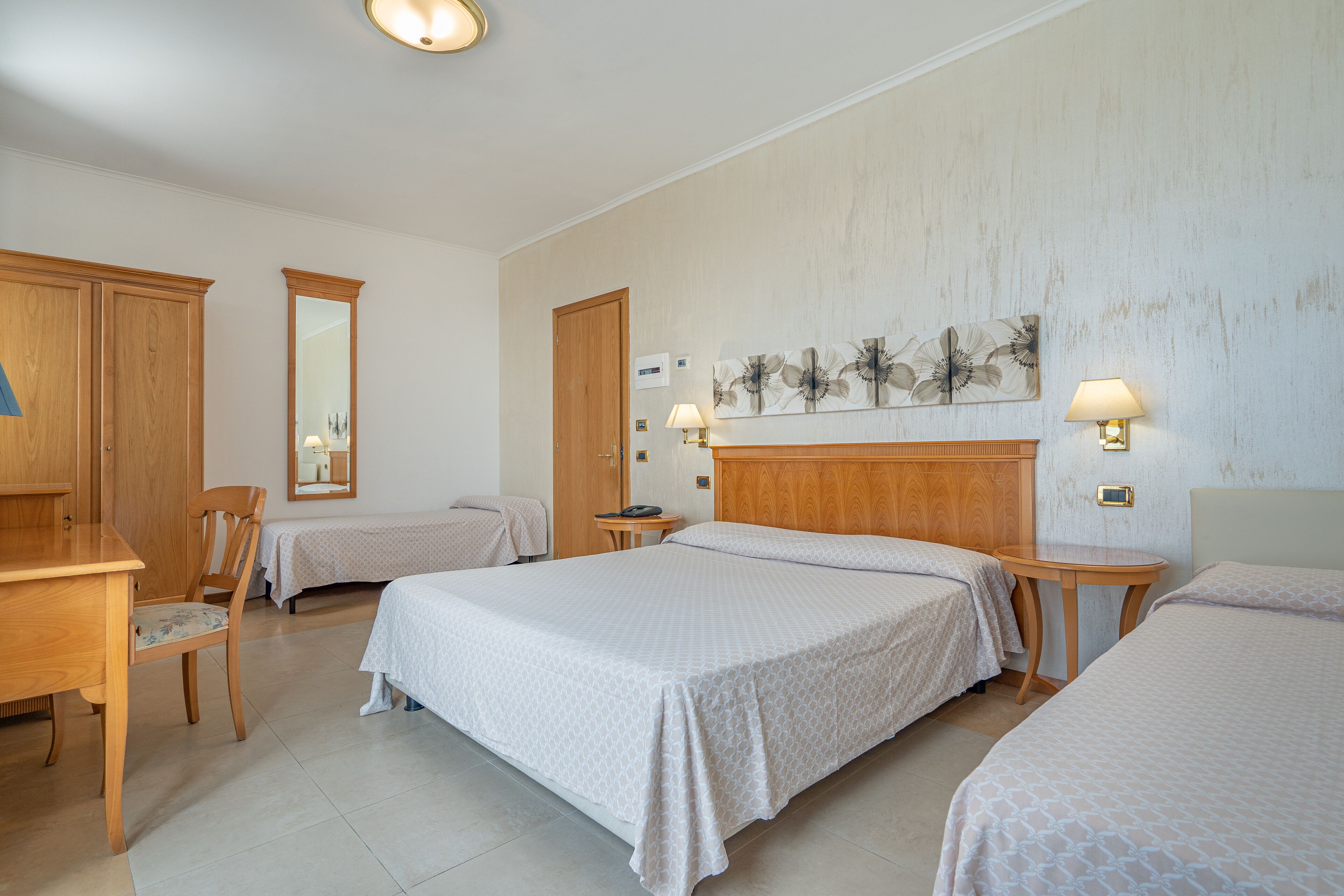 A hotel room with two beds, a wooden desk, a chair, a mirror, and a wall-mounted lamp.