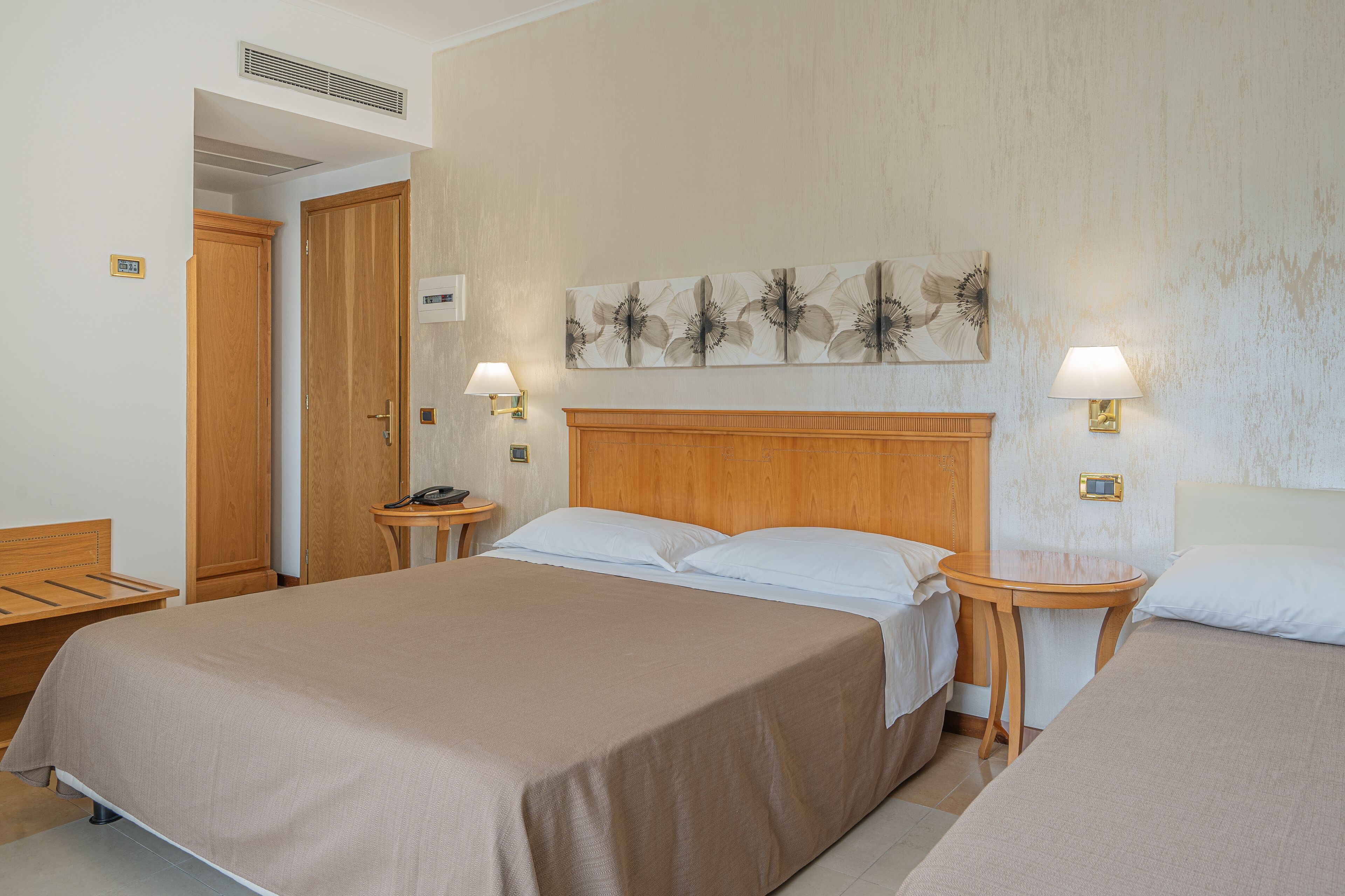 Une chambre d’hôtel avec un grand lit, deux tables de chevet, une tête de lit en bois, une œuvre d’art accrochée au mur et une porte donnant sur une autre pièce.