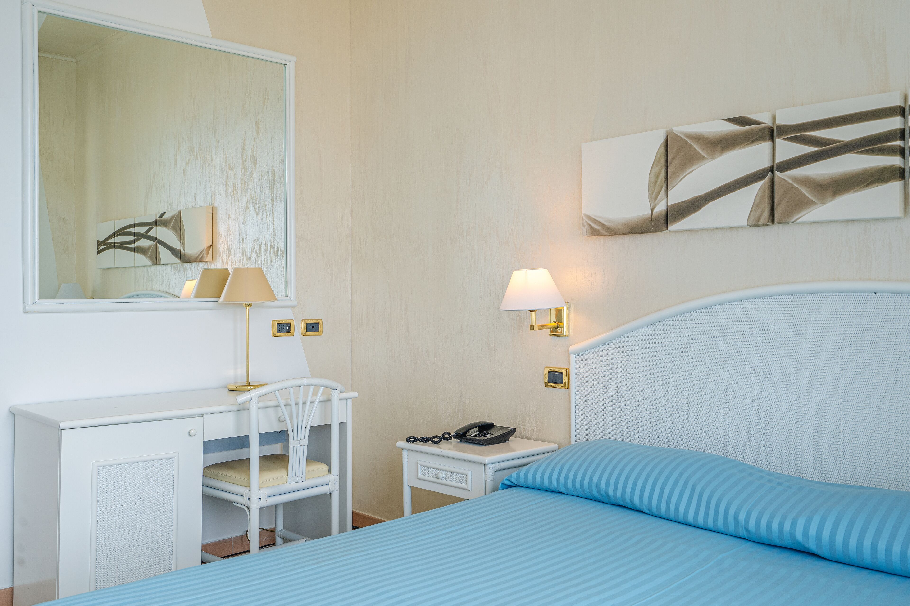 A hotel room with a bed, a desk, a chair, a bedside table with a lamp, and artwork on the walls.