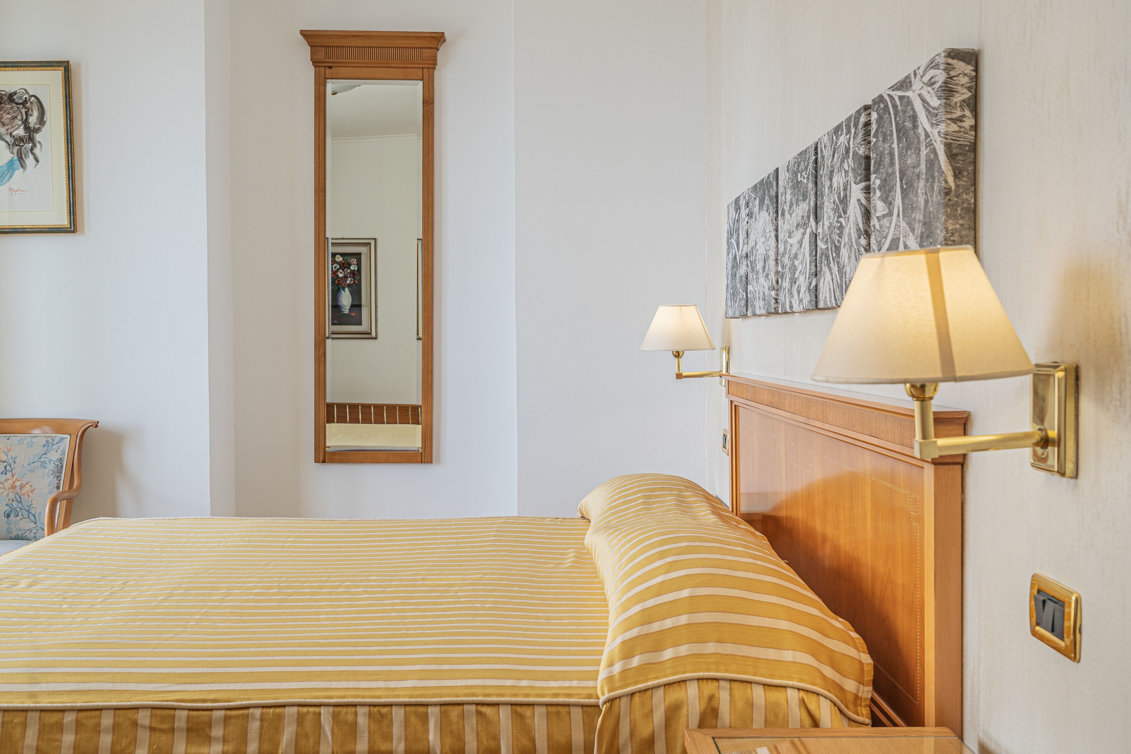 A neatly made bed with striped bedding, a wooden headboard, a wall-mounted mirror, and a framed picture.