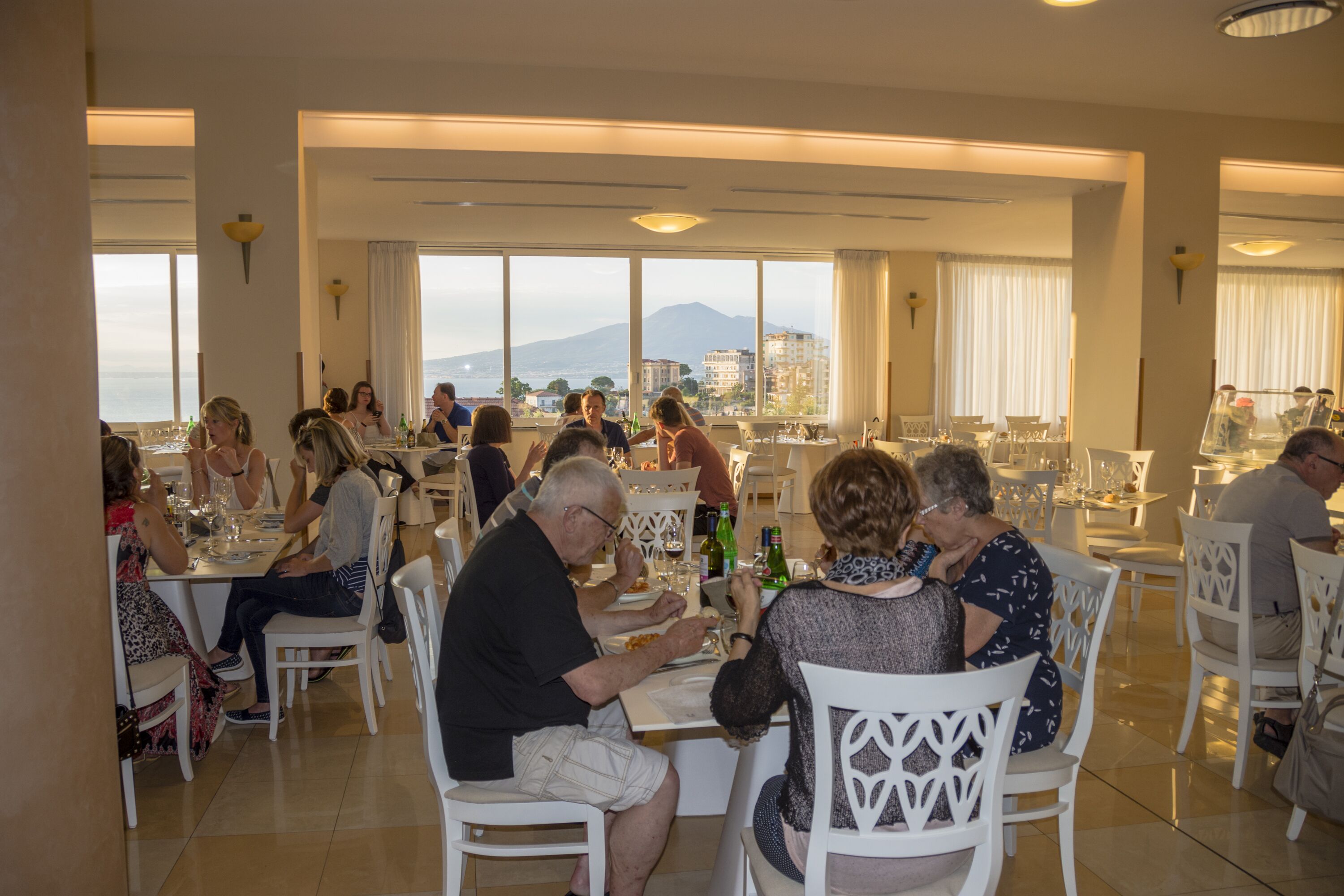 Um restaurante com cadeiras e mesas brancas, amplas janelas com vista para o mar e montanhas, e um ambiente acolhedor para refeições.