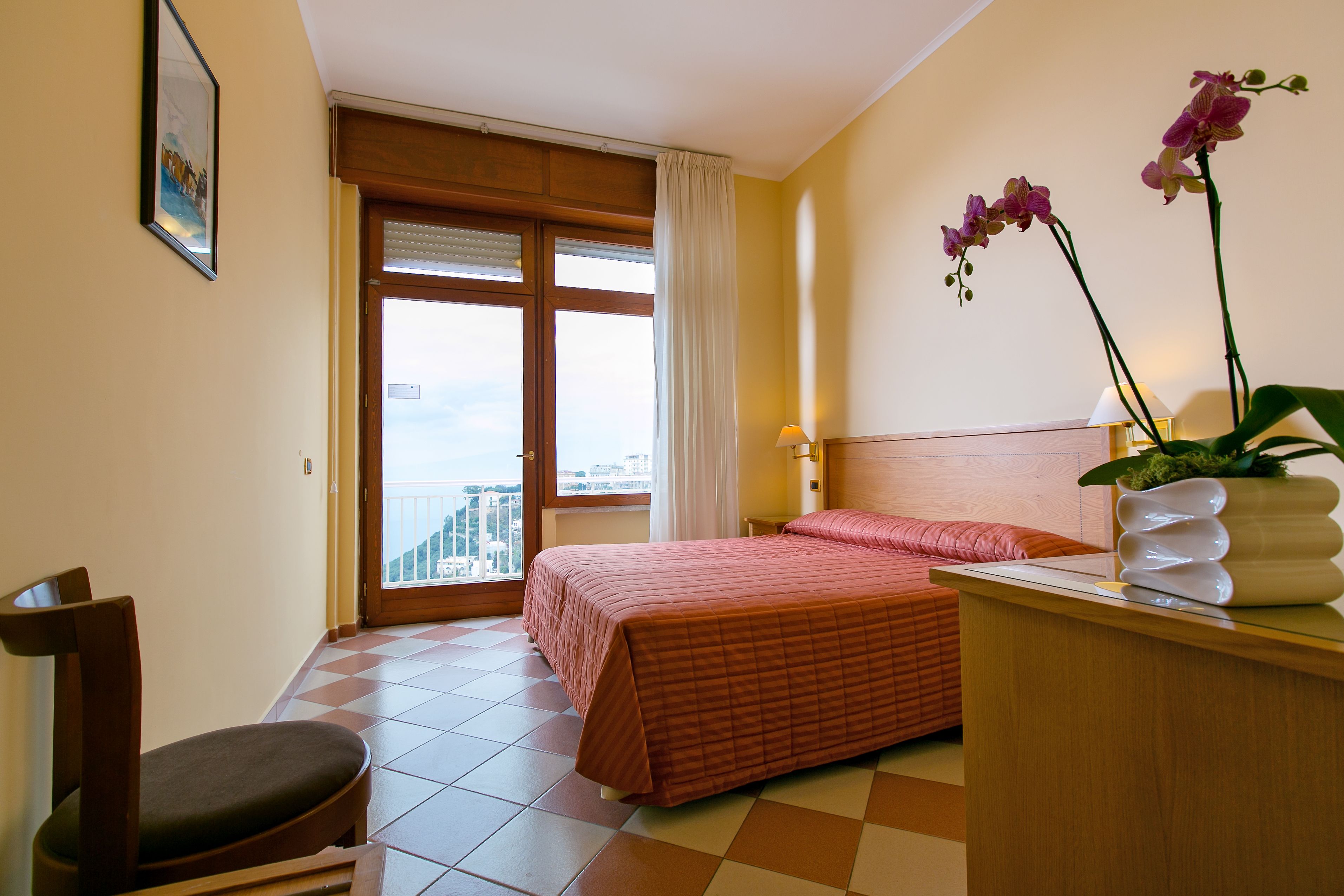 Quarto de hotel com cama, uma cadeira, escrivaninha com um vaso de flores e janela com vista.