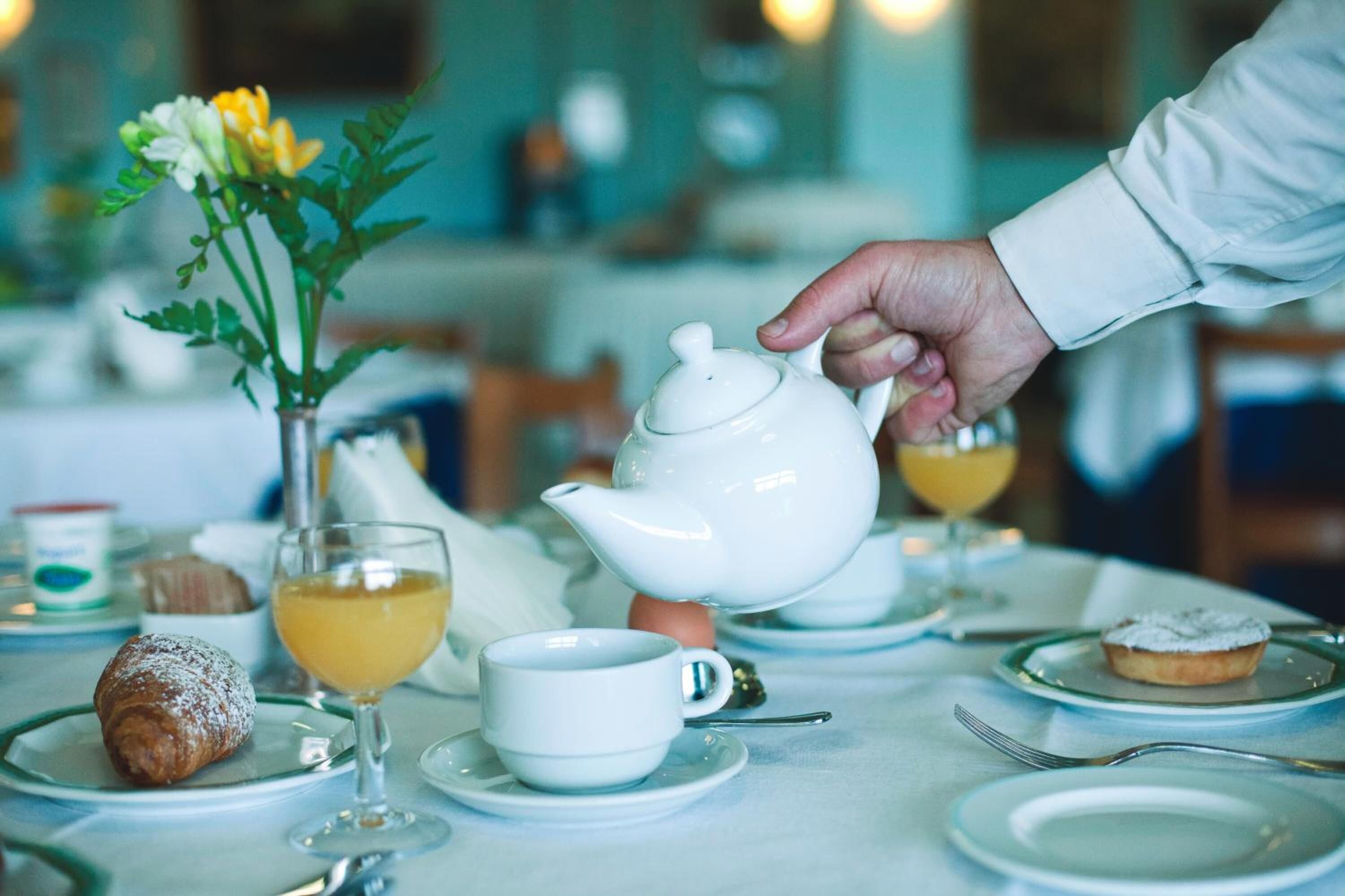 Ein Esstisch mit einem Teekessel, einer Tasse, einem Glas Orangensaft, einem Teller mit Gebäck und einer Vase mit Blumen.