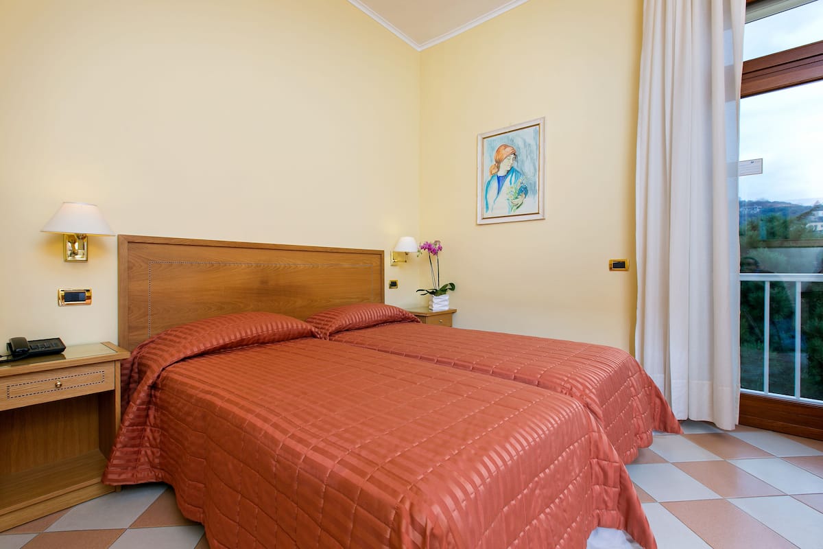 A hotel room with a large bed, two bedside tables, a nightstand, a painting on the wall, and a window with curtains.