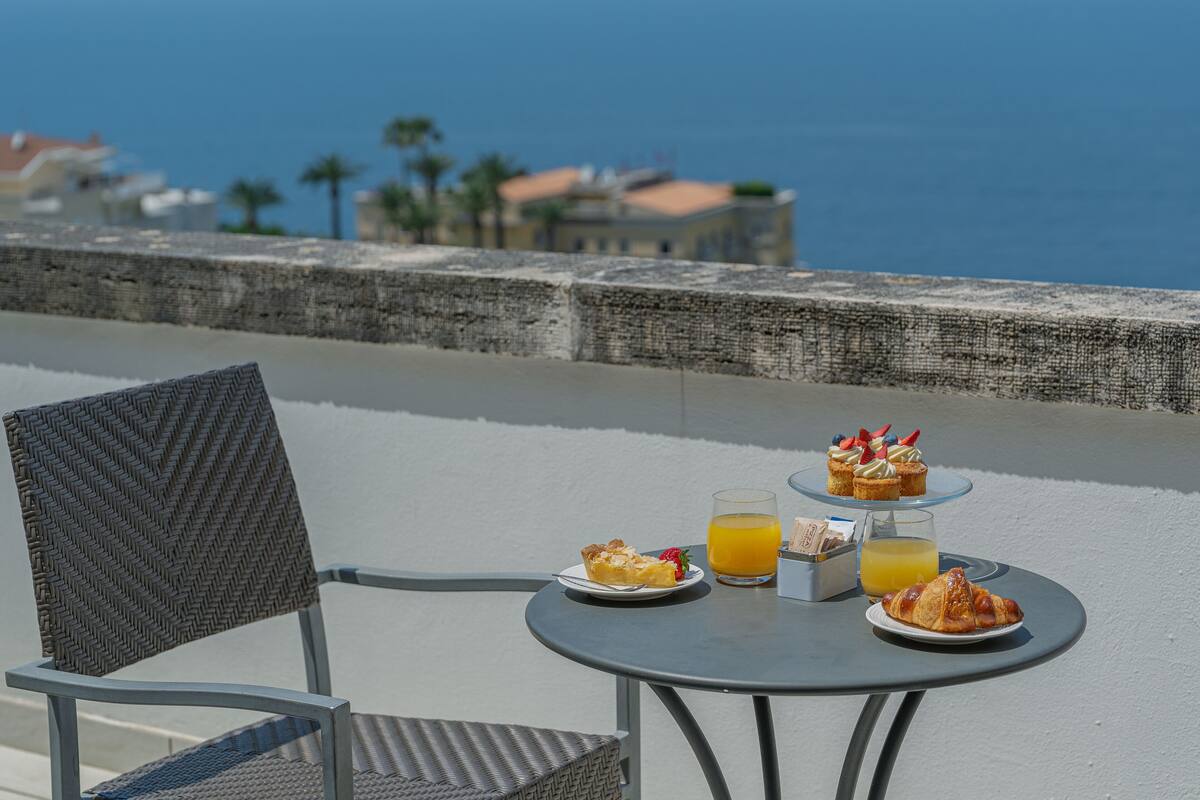 A table with breakfast items including pastries, a glass of orange juice, and a plate of food, set on a balcony overlooking a coastal area.