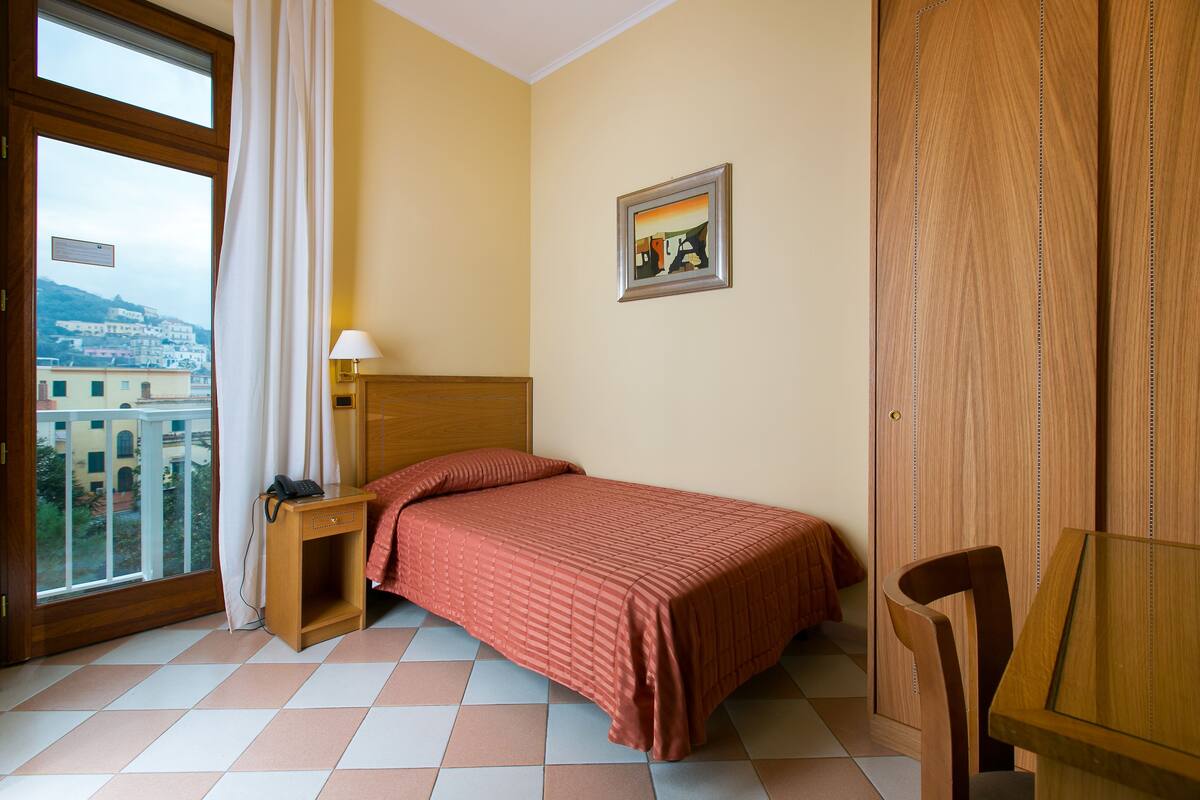 A bedroom with a bed, a nightstand, a desk, and a chair. There is a window with a view of buildings and a door.
