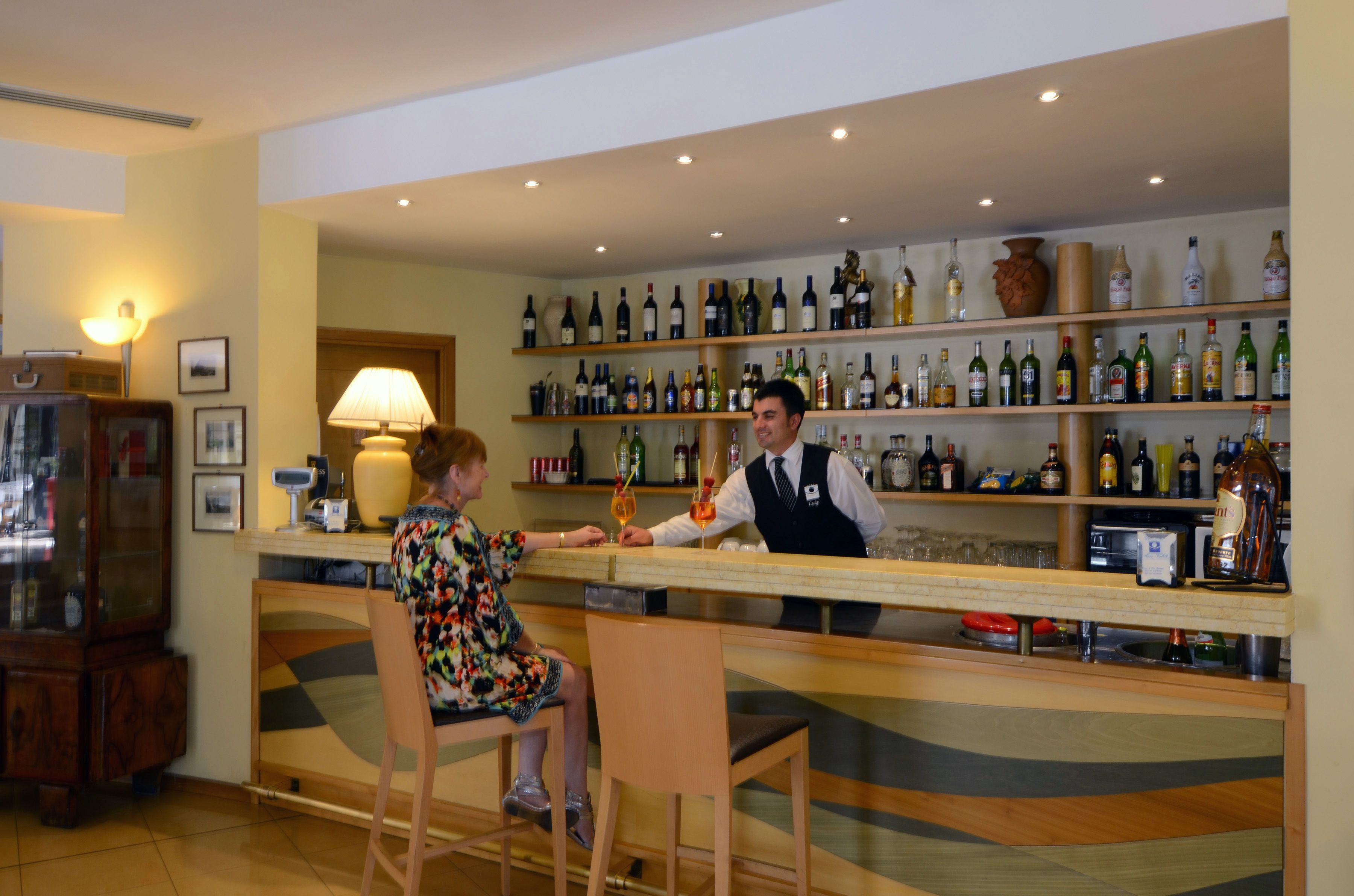 A bar area with a bartender and a customer. The bar has shelves with various bottles and glasses. There is a lamp on the wall and a cabinet with glass doors.