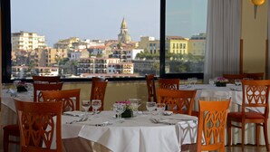 A dining area with tables set for six, each with white tablecloths and wooden chairs. A large window offers a view of a cityscape with buildings and a prominent tower. - Grand Hotel Moon Valley (Vico Equense)