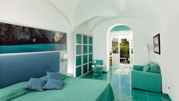 A room with a bed, a chair, a sofa, a painting of a sea, and a view of the sea through an archway. - Gatto Bianco Hotel & SPA (Capri)