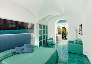 A room with a bed, a chair, a sofa, a painting of a sea, and a view of the sea through an archway.