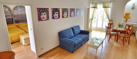 A living room with a blue sofa, a glass coffee table, and a dining area with wooden chairs and a table. There are four paintings on the wall and a door leading to another room.