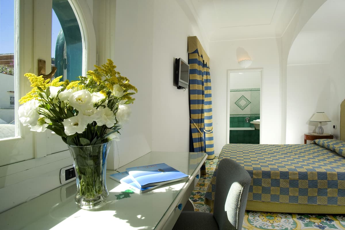 A hotel room with a bed, a desk with a vase of flowers, a television, and a bathroom visible through an arched doorway.