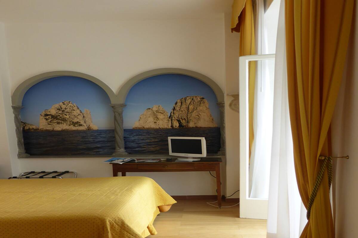 A bedroom with a bed, a desk with a computer, and a mural of a coastal scene.