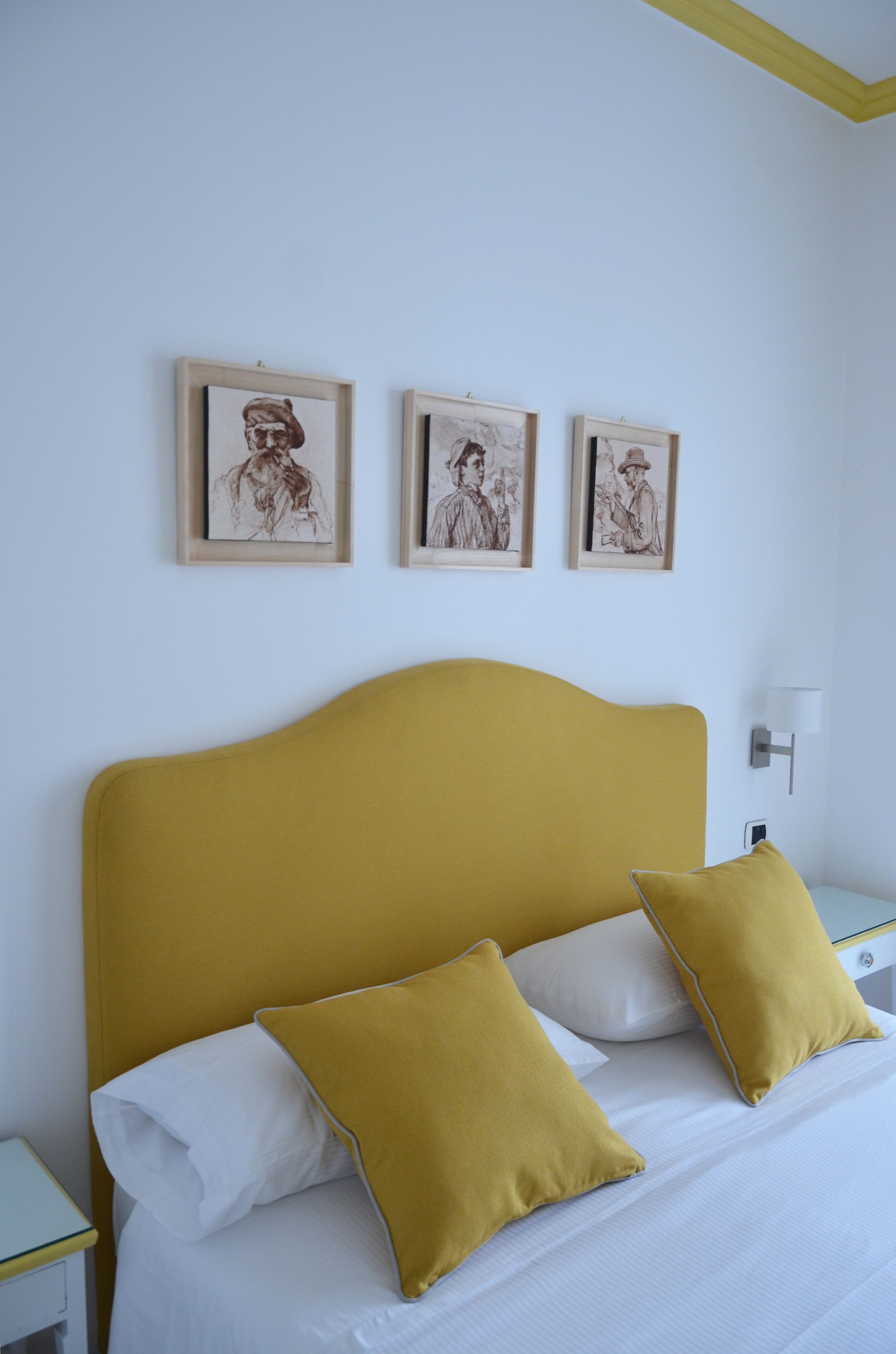 Una cama con sábanas blancas y almohadas color mostaza, un cabecero color mostaza y tres dibujos enmarcados en la pared de arriba.