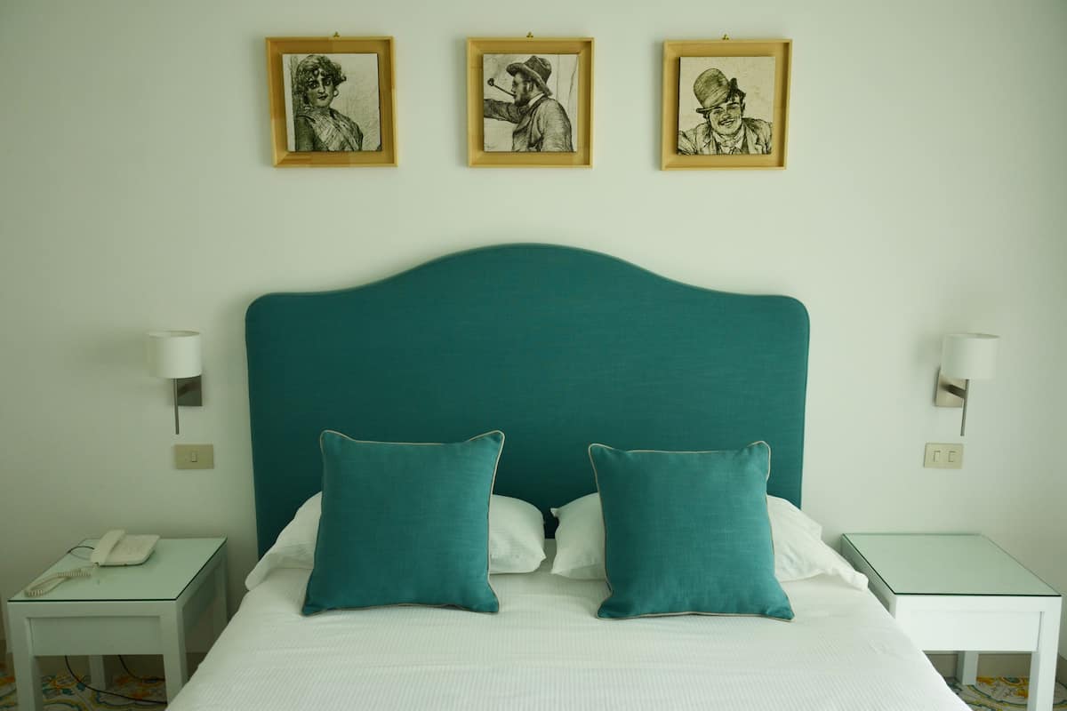 A bed with teal headboard, white bedding, and teal pillows, flanked by bedside tables with lamps and a phone.
