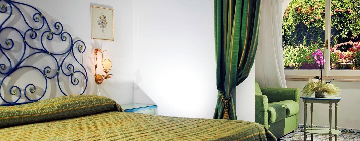 A bedroom with a bed, a green armchair, a small table, a wall-mounted light fixture, and a window with a view of greenery.
