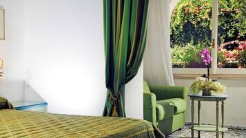 A bedroom with a bed, a green armchair, a small table, a wall-mounted light fixture, and a window with a view of greenery.