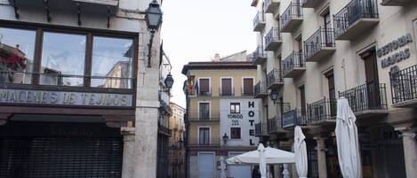 Una calle con edificios, incluyendo un hotel y una tienda de textiles. Hay mesas y sombrillas al aire libre.