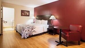 Habitación de hotel con cama, lámparas de noche, un sillón rojo, una mesita y una puerta que da a un baño.