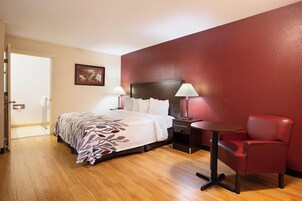Habitación de hotel con cama, lámparas de noche, un sillón rojo, una mesita y una puerta que da a un baño.