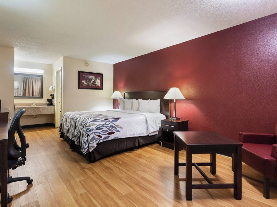 A hotel room with a bed, bedside lamps, a desk, a chair, and a red accent wall.