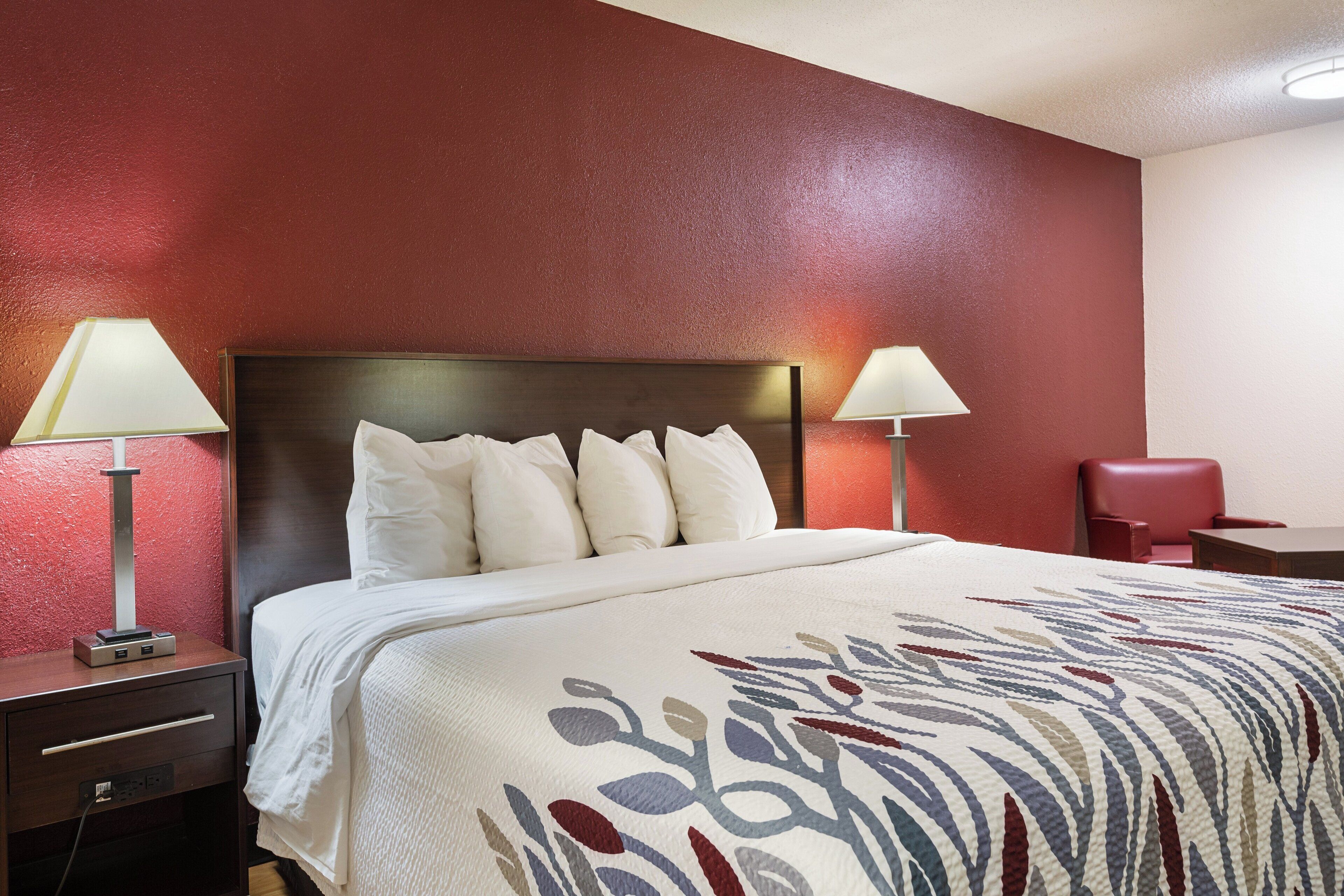 A hotel room with a large bed, two bedside lamps, a red wall, and a small desk with a chair.