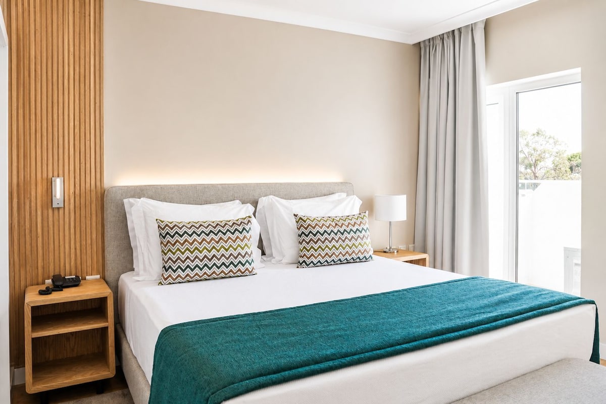 A hotel room with a bed, two pillows, a nightstand, a lamp, and a view of the outdoors.